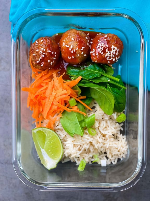 Three meatballs with sauce and sesame seeds, shredded carrot, spinach, rice, and lime wedg