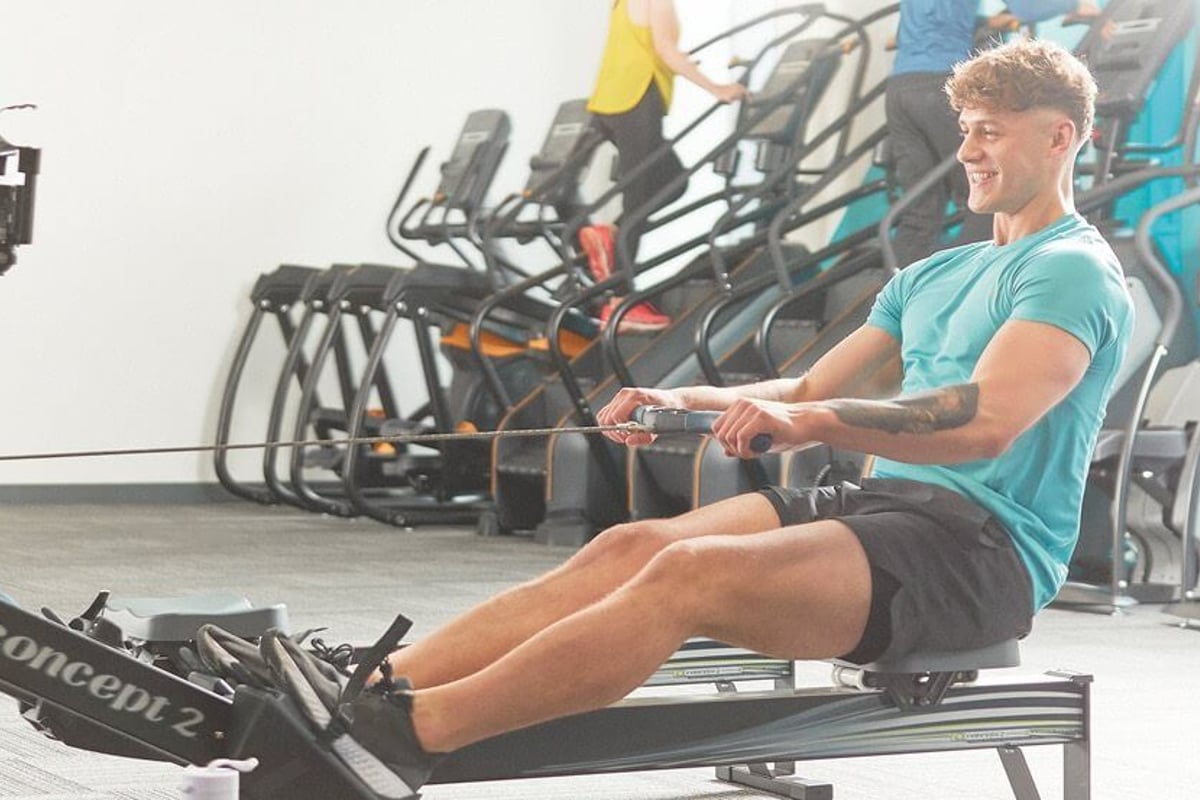Man in turquoise shirt using rowing machine in gym with treadmills in background