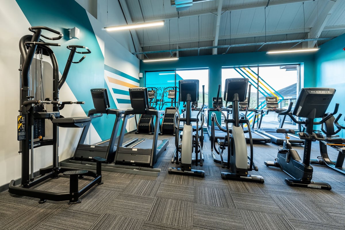 PureGym Kendal: Treadmills, stationary bikes, weight machines, patterned carpet flooring in a vibrant blue fitness studio.