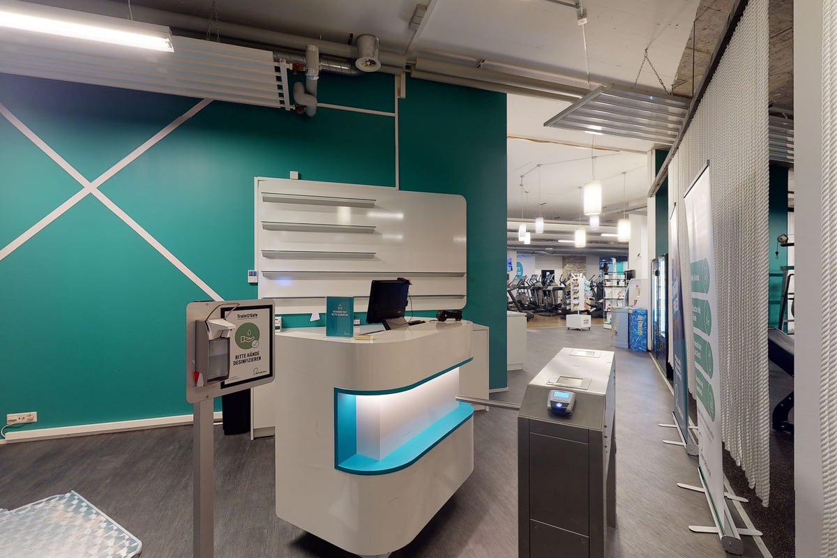 PureGym Chur: Empfangsbereich, Fitnessgeräte, die durch Glastrennwände sichtbar sind, zeitgenössisches Design mit blaugrünen Akzenten.