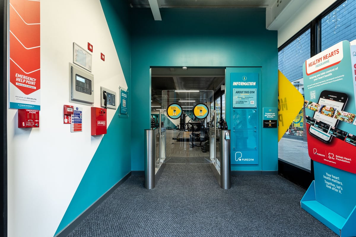 PureGym Droitwich Spa: Teal corridor with turnstiles, signage, and information kiosks.