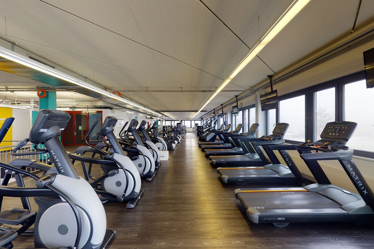 PureGym Uster: Des rangées de tapis de course bordent l'intérieur d'une salle de sport bien éclairée avec des fenêtres allant du sol au plafond le long d'un mur.
