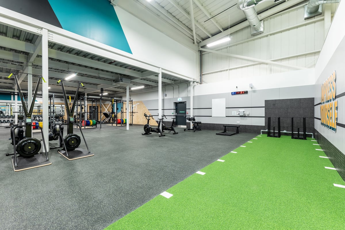 PureGym Eastbourne: Weightlifting rigs, barbells, kettlebells, sleds on rubberised flooring alongside green turf area.
