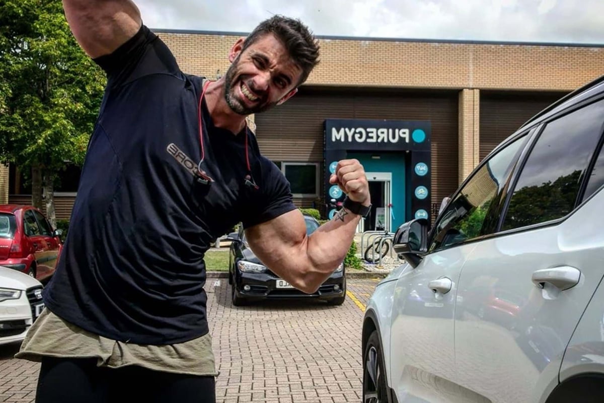 muscular man flexing arms in front of gym building and parked cars on cloudy day