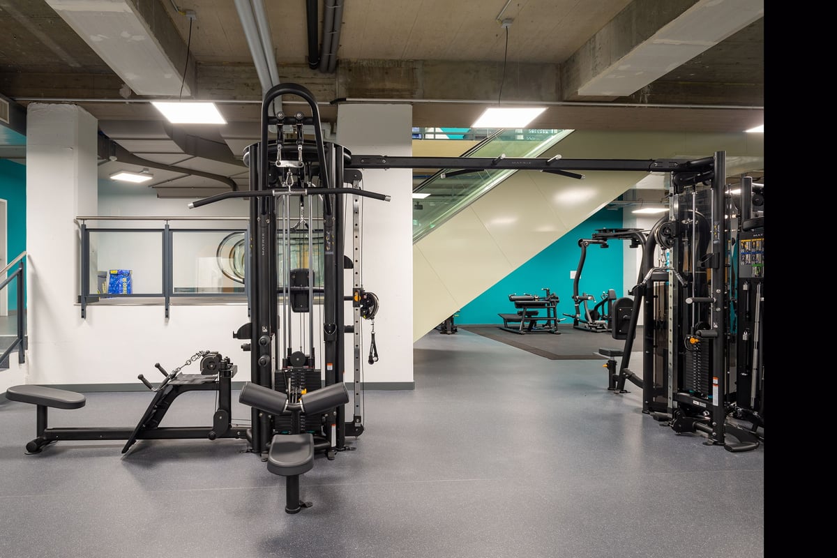 PureGym La Chaux-de-fonds: Gym studio with weight machines, benches, and teal accents against white walls and floors.