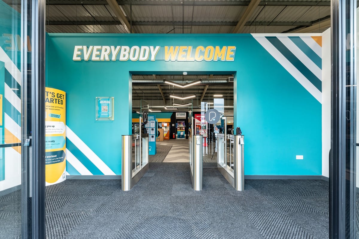PureGym Merthyr Tydfil: Entrance corridor with teal walls, turnstiles, and directional signage welcoming visitors.