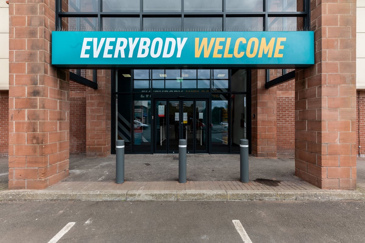 PureGym Dumfries: Brick building entrance with glass doors and an "EVERYBODY WELCOME" sign above the doorway.