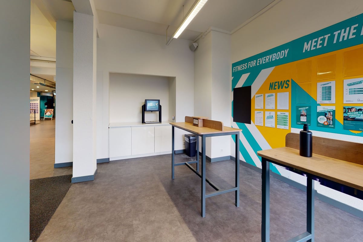 PureGym Bern Lorraine: Hall d'entrée avec tables, chaises, kiosque numérique et signalisation favorisant l'accès à l'information pour tous.