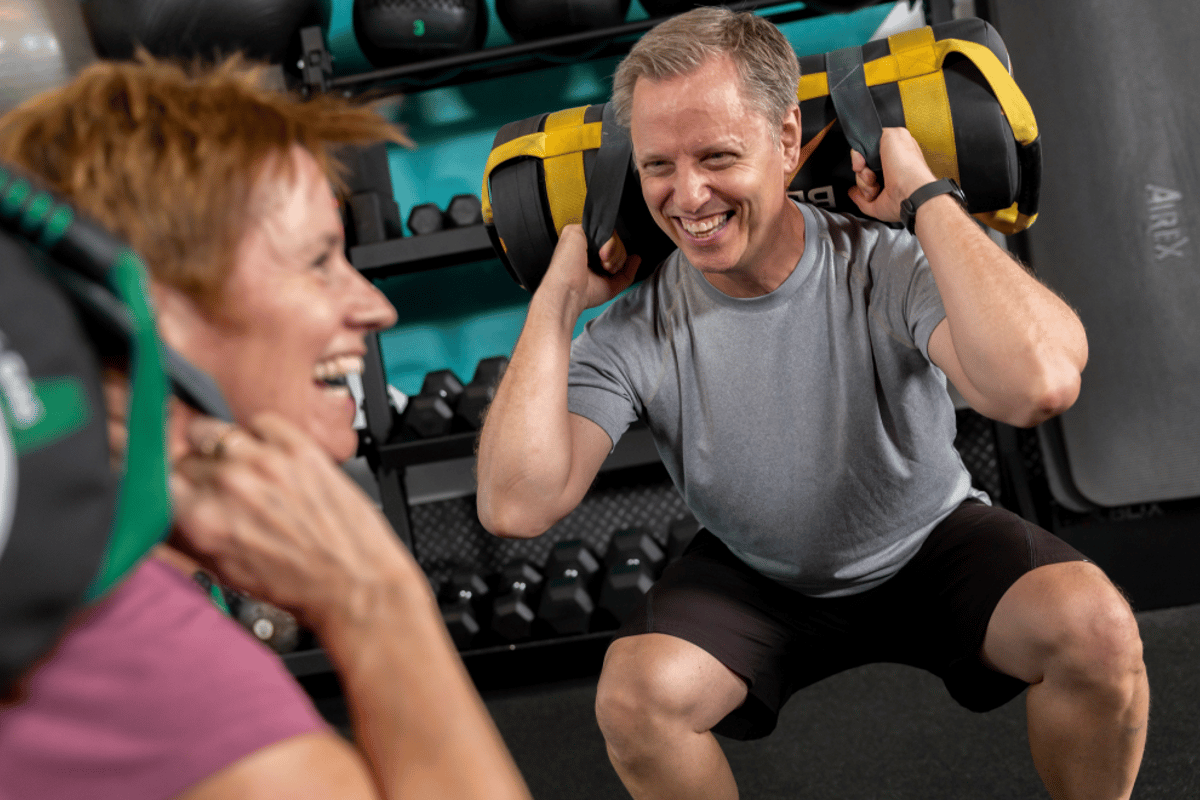 Zwei Personen lächeln beim Kniebeugen mit Gewichtssäcken auf den Schultern im Fitnessstudi