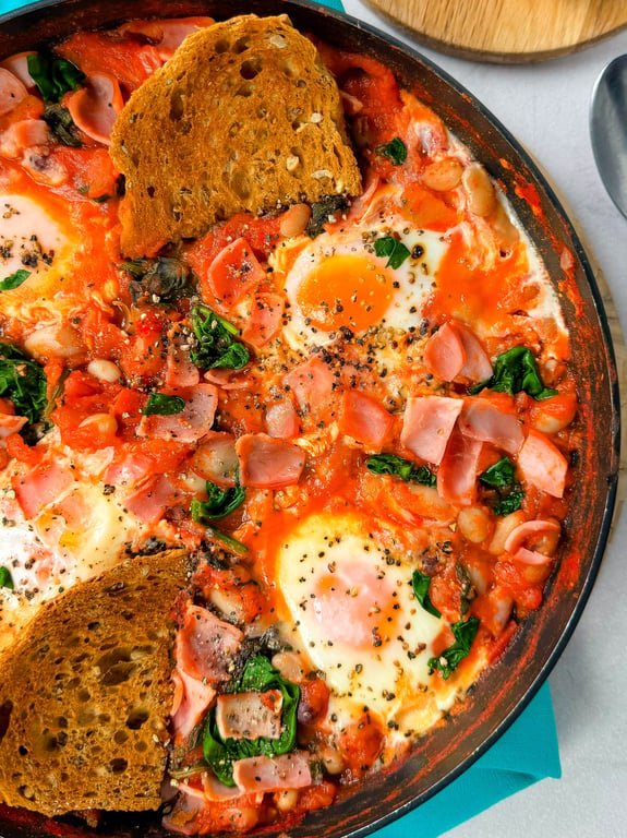 Shakshuka with eggs, spinach, ham, beans, tomato sauce, and toasted bread slices in pan