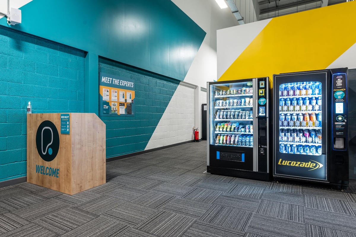 PureGym Alton: Vending machines, welcome desk, tiled walls in teal and yellow colour scheme.