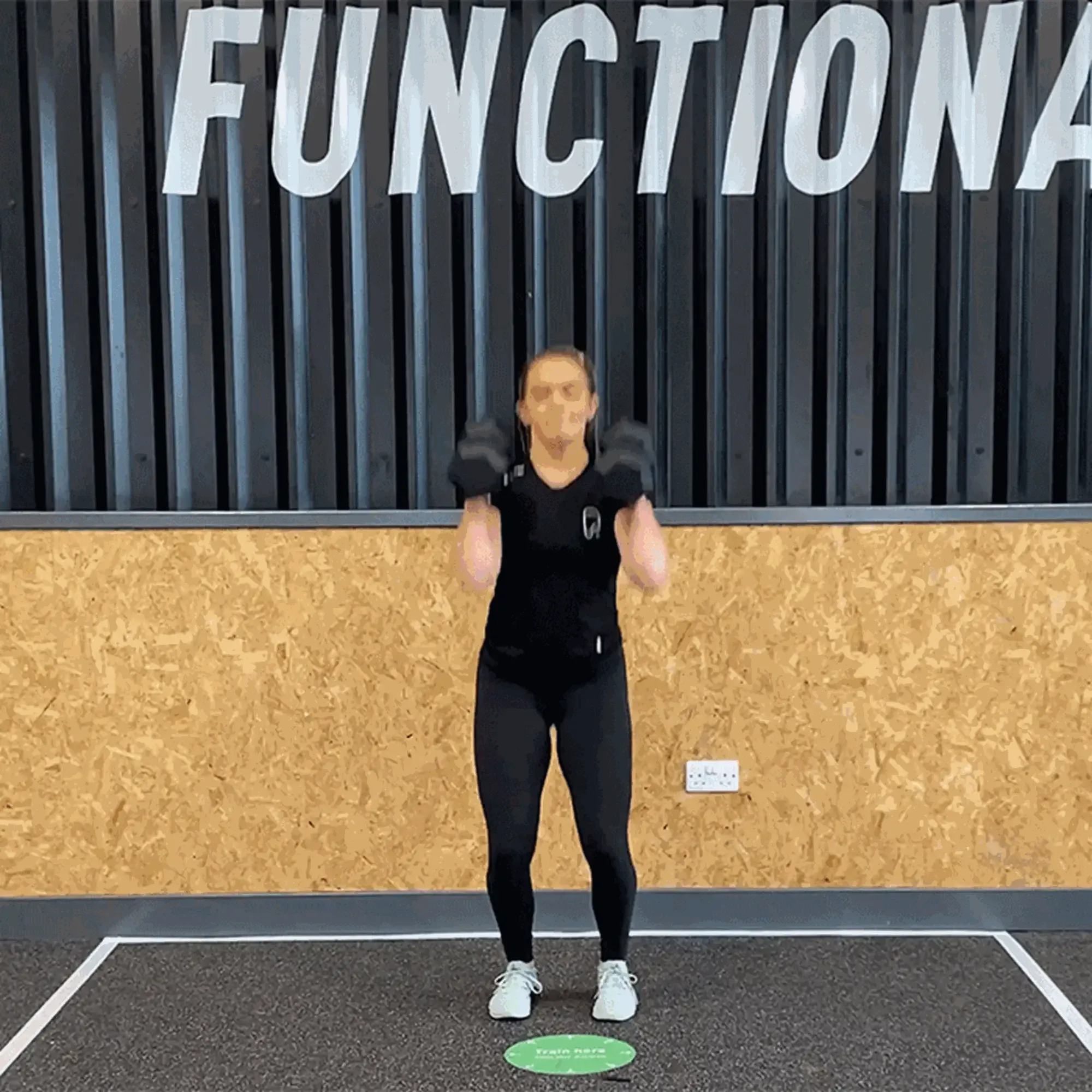 Woman in black workout clothes lifting dumbbells in a gym with FUNCTIONAL sign above