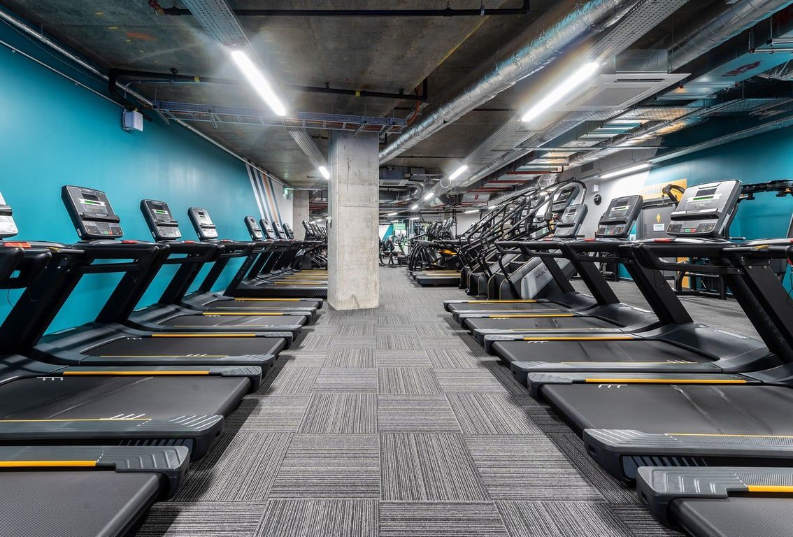 PureGym London Hoxton: Rows of modern treadmills in a dimly lit gym interior with teal walls and concrete pillars.