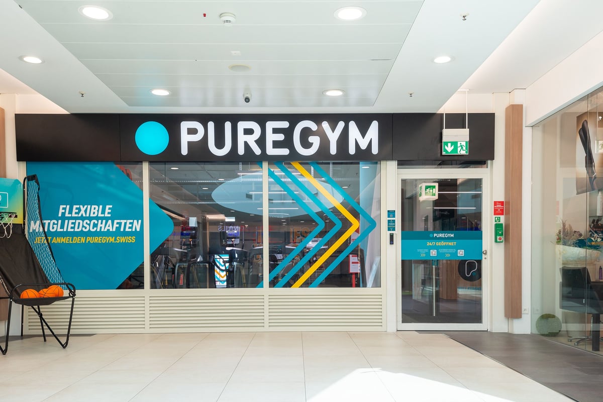 PureGym Wil: Vitrine Pure Gym avec entrée vitrée, signalisation et flèches indiquant les adhésions flexibles.