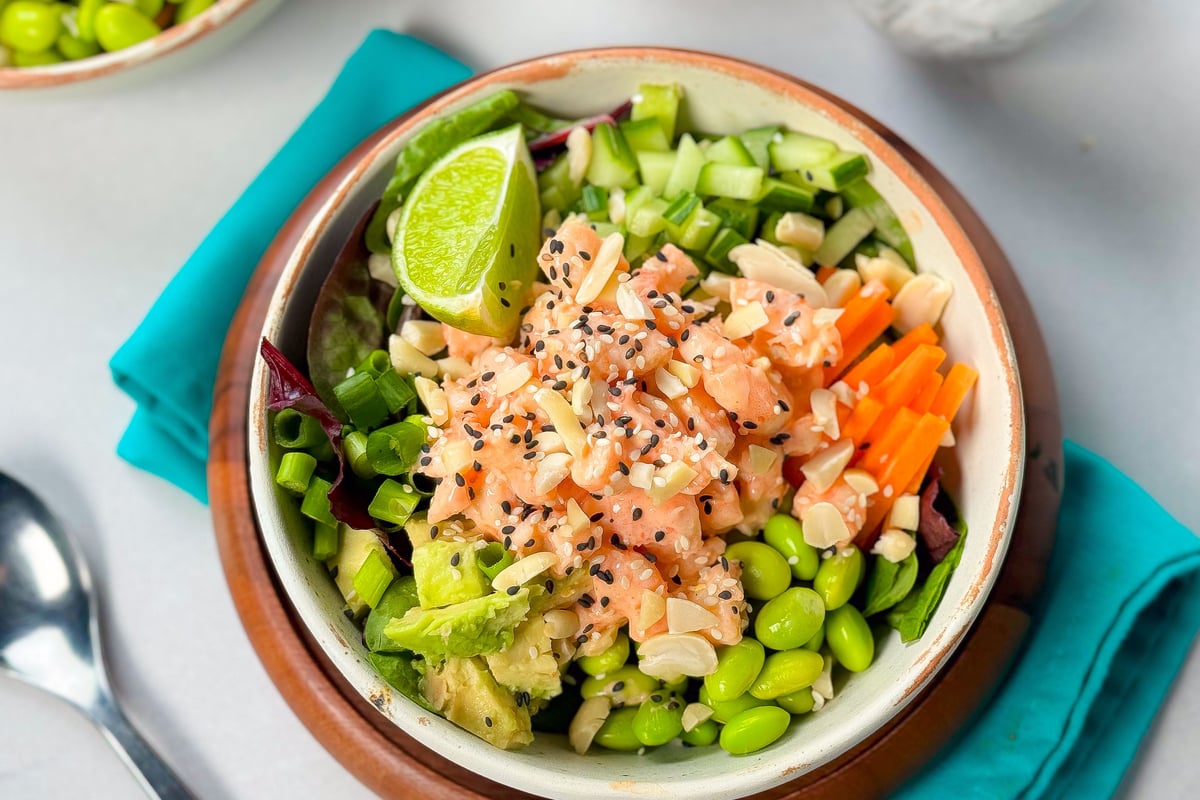 Bowl of prawn poke with edamame, cucumber, carrot, avocado and lime on turquoise napkin