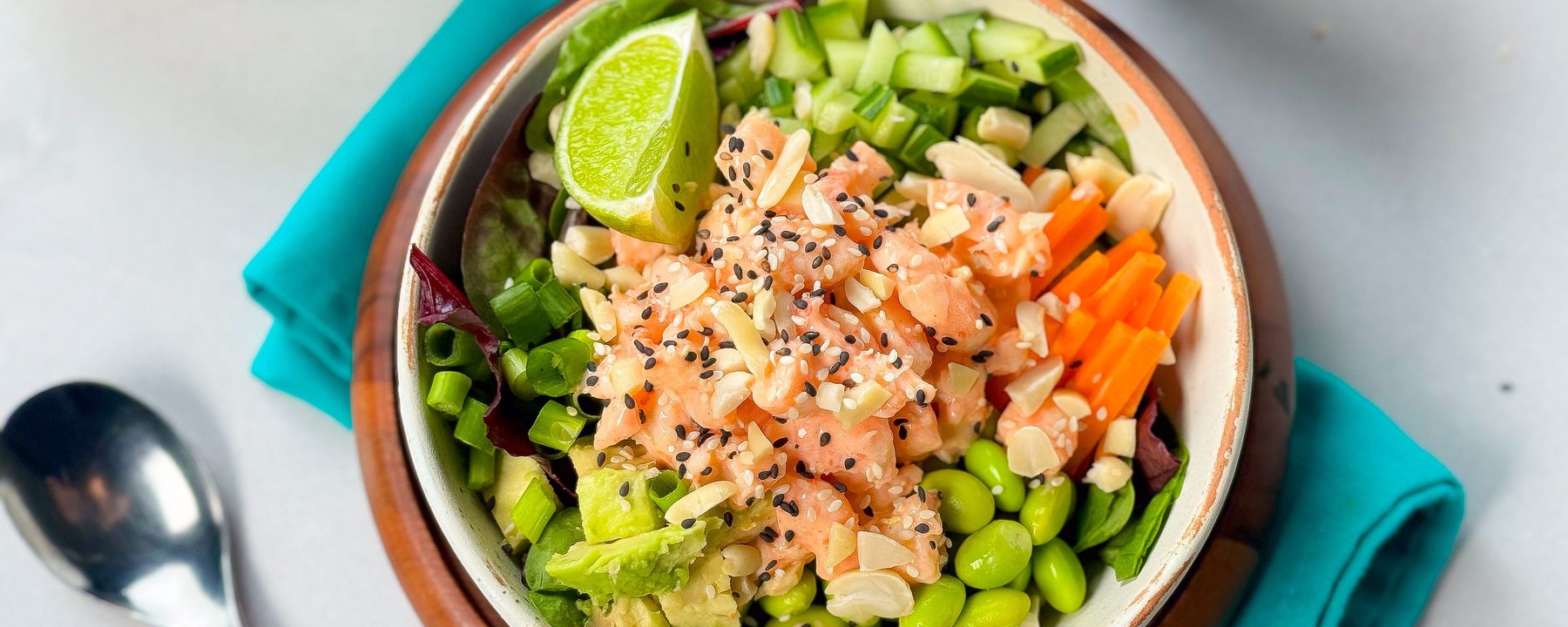Bowl of prawn poke with edamame, cucumber, carrot, avocado and lime on turquoise napkin