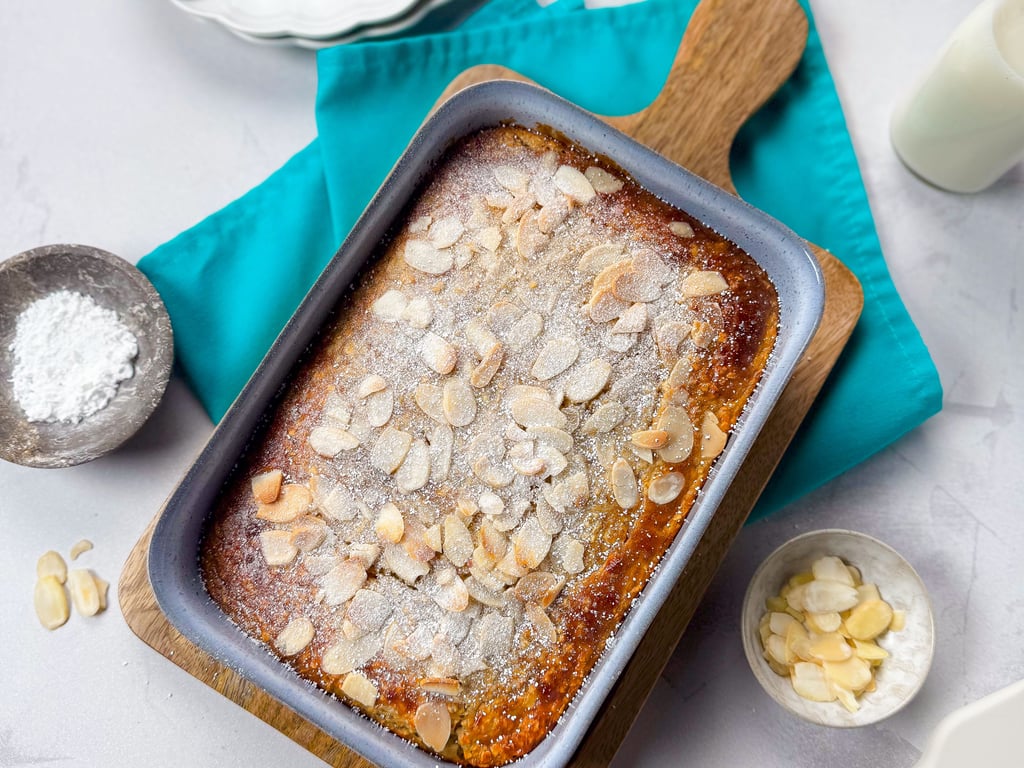 Baked almond cake in a blue tray on a wooden board with powdered sugar and almonds nearby