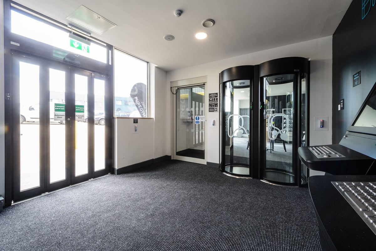PureGym Billericay: Revolving doors, exercise bicycles behind glass, emergency exit signs.