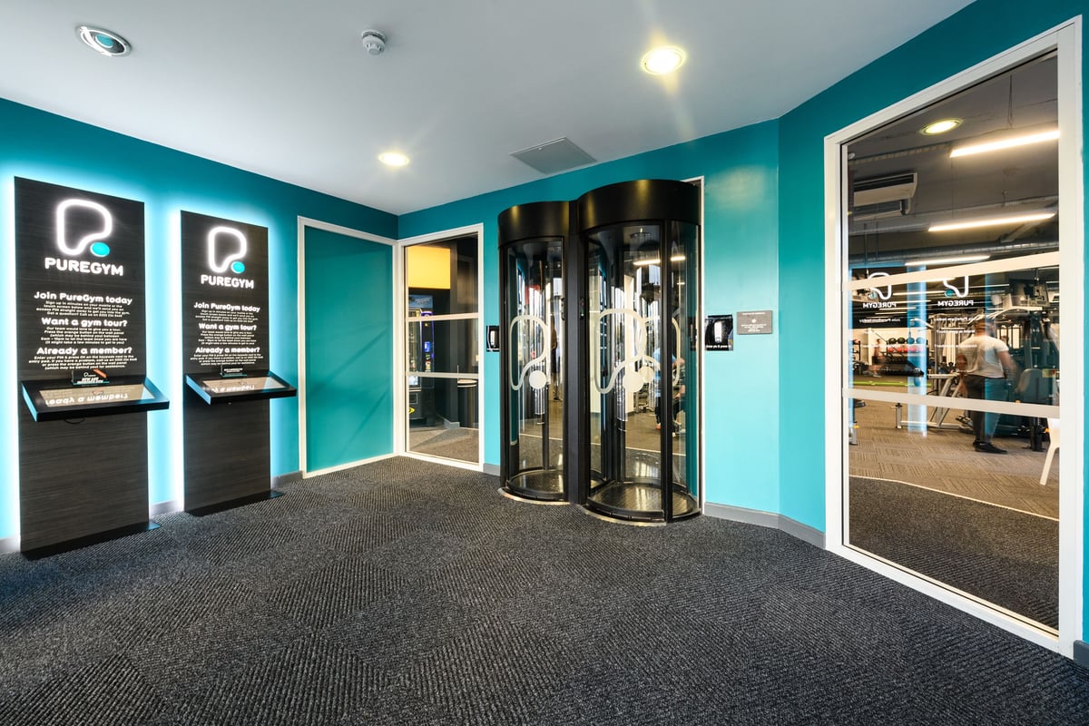 PureGym Bournemouth The Triangle: Revolving doors, fitness kiosks, teal decor, and carpeted gym entrance area.