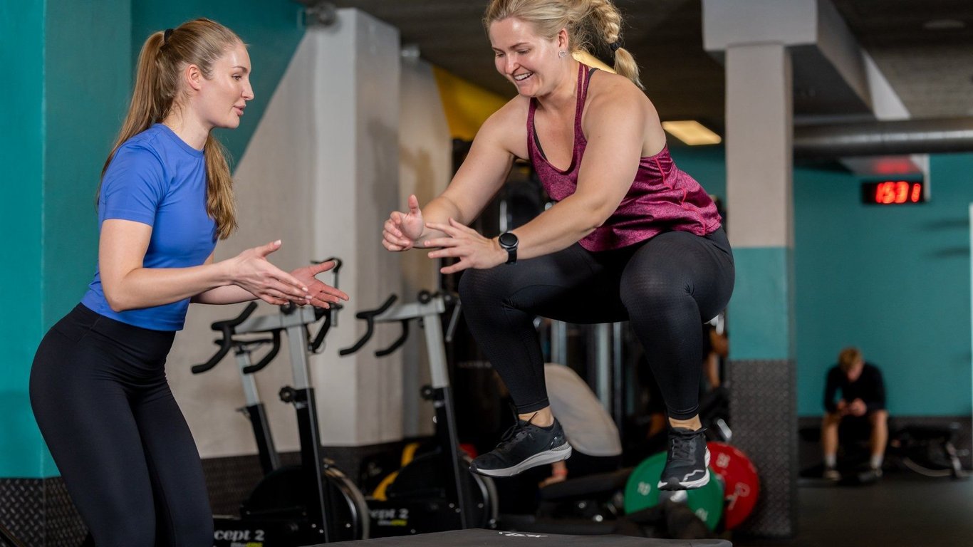 Woman in purple top performing box jump exercise whilst trainer in blue top observes