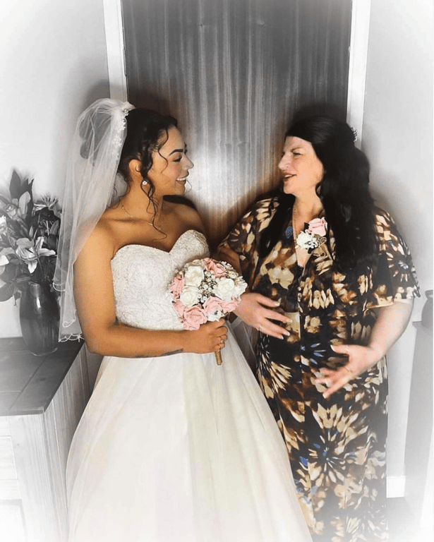 Bride in white dress holding bouquet talking to woman in patterned dress indoors