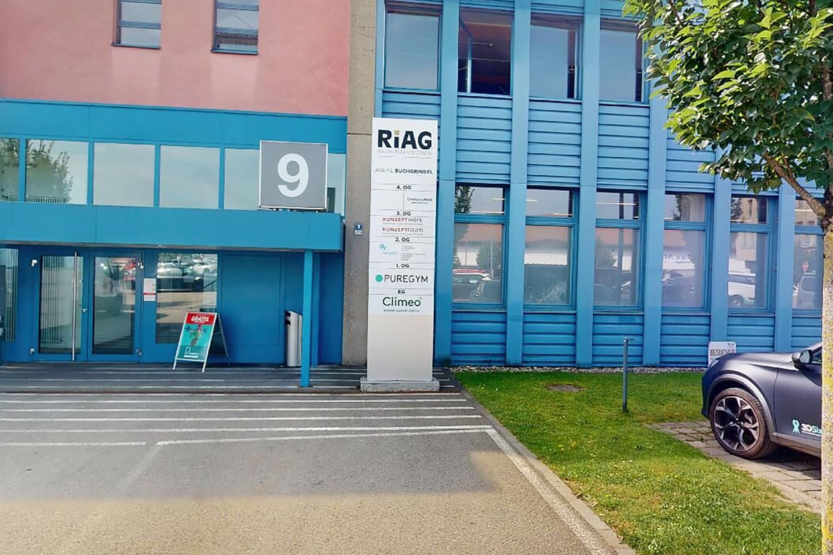 PureGym Wetzikon: Aussenansicht des Riag-Gebäudes mit Glaseingang, draussen geparktem Auto und grünem Grasstreifen.