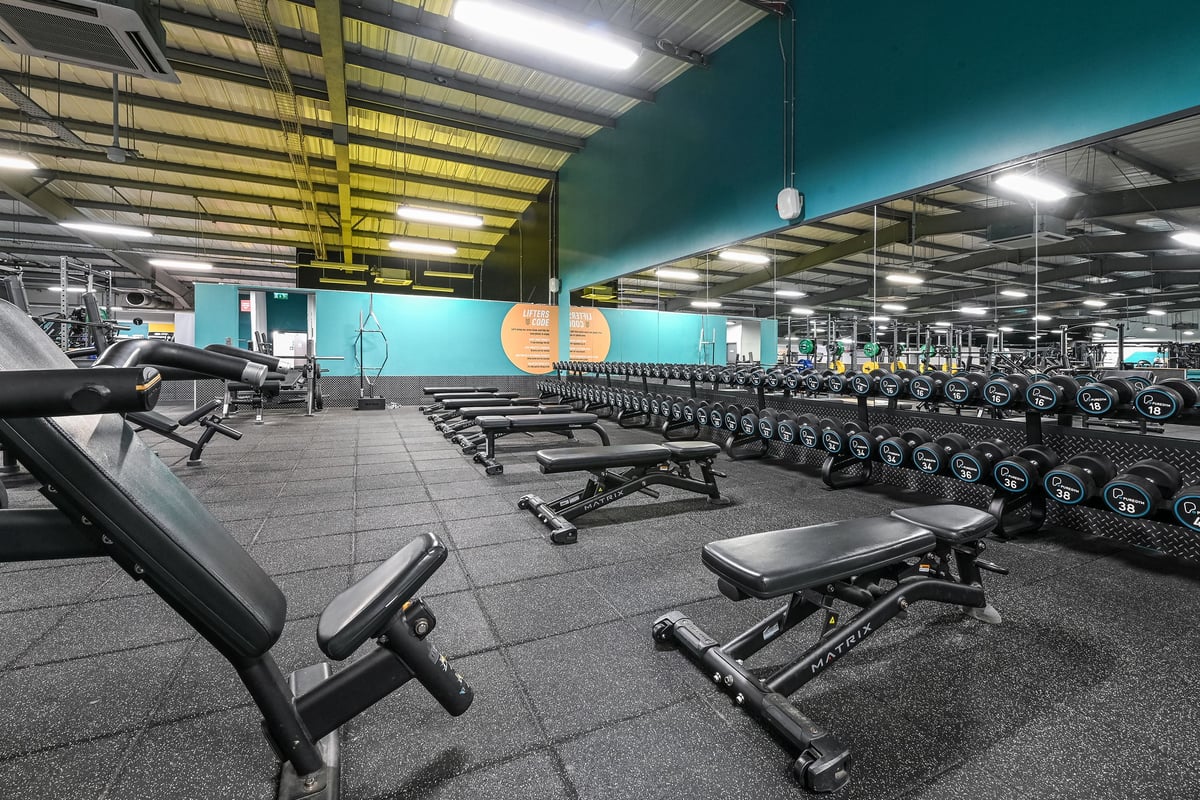 PureGym Canterbury Sturry Road: Rows of weight benches, dumbbells, and squat racks on rubberized flooring in a gym facility.