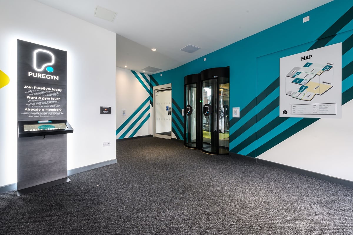 PureGym London Fulham: Gym entry with turnstiles, teal accent walls, and digital screens displaying map.