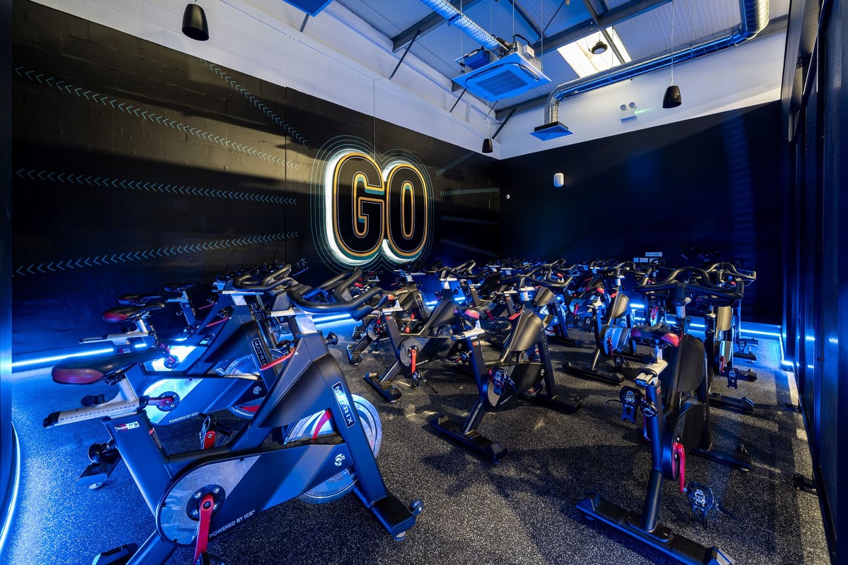 PureGym Newtownabbey: Rows of stationary cycles in dimly lit studio with neon 'GO' sign and blue lighting.