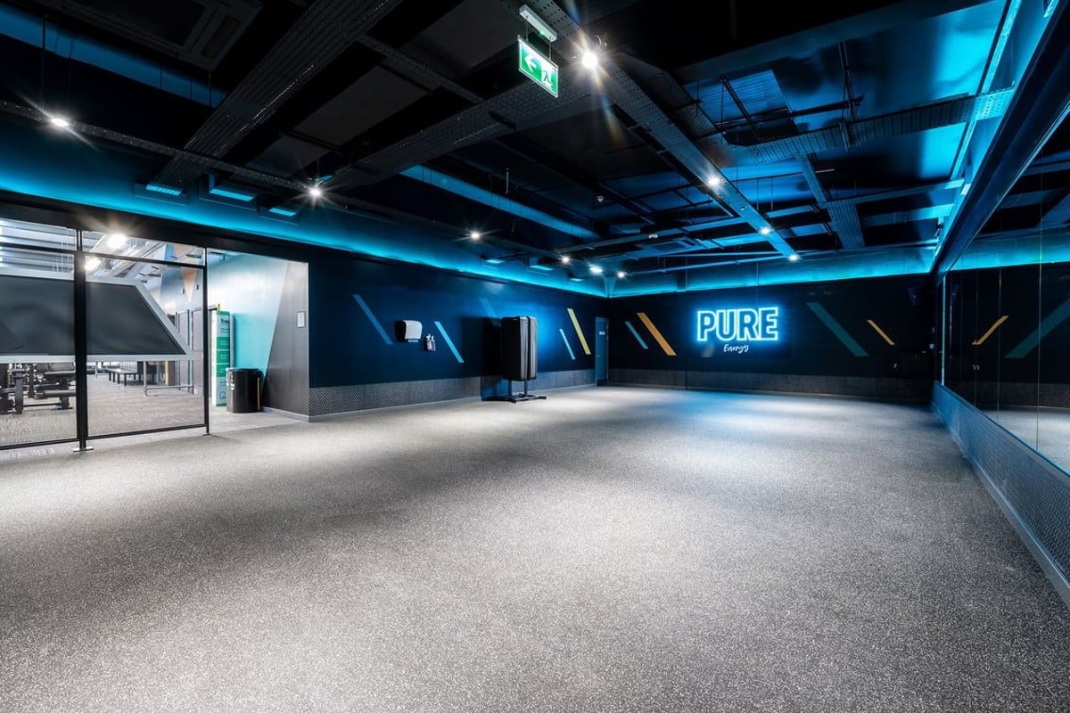 PureGym Inverness Inshes Retail Park: Bare gym floor, teal lighting, exposed ceiling with signage and exit sign visible.