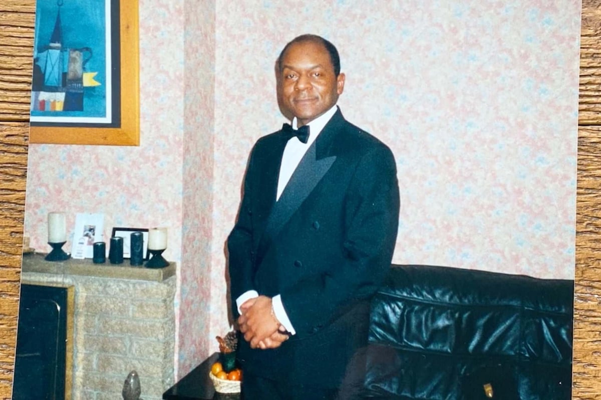 Man in black tuxedo standing in a living room with a leather sofa and framed art on wall