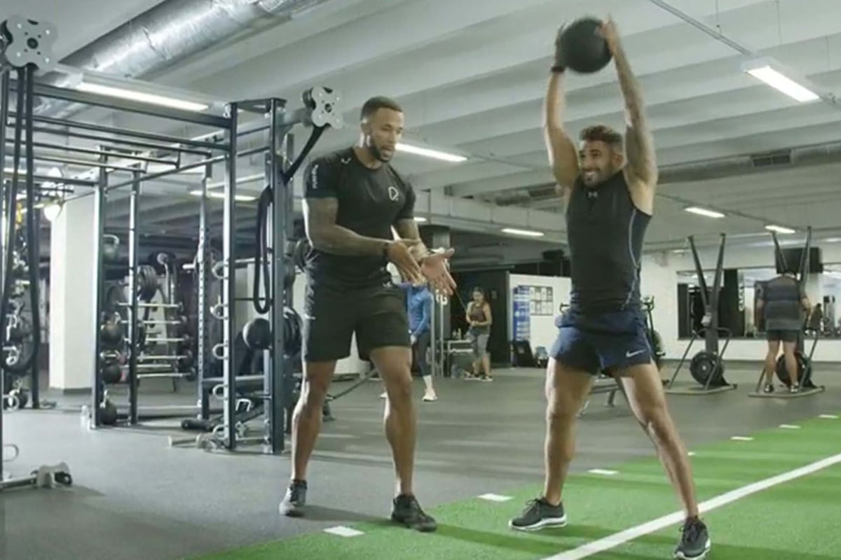 Man lifting medicine ball overhead while trainer watches in gym with equipment