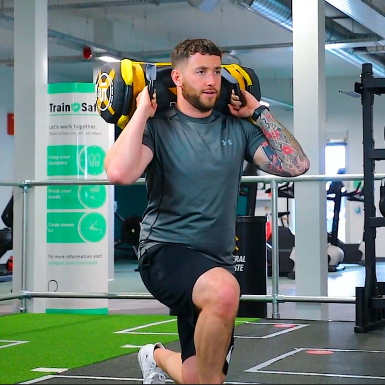 Man with tattooed arm doing weighted lunges in gym with safety sign behind him