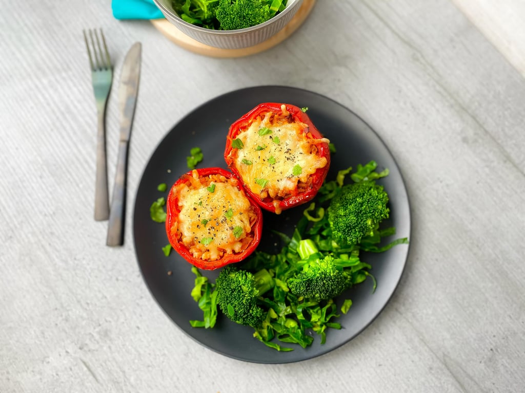 Two stuffed red peppers with melted cheese and herbs on a black plate with broccoli