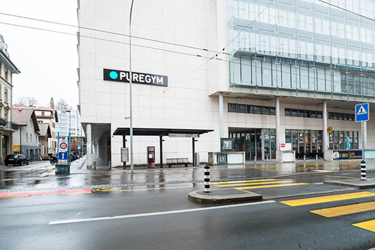 PureGym Fribourg: Rue pluvieuse avec entrée du gymnase, marquage des passages pour piétons et signalisation commerciale visibles.