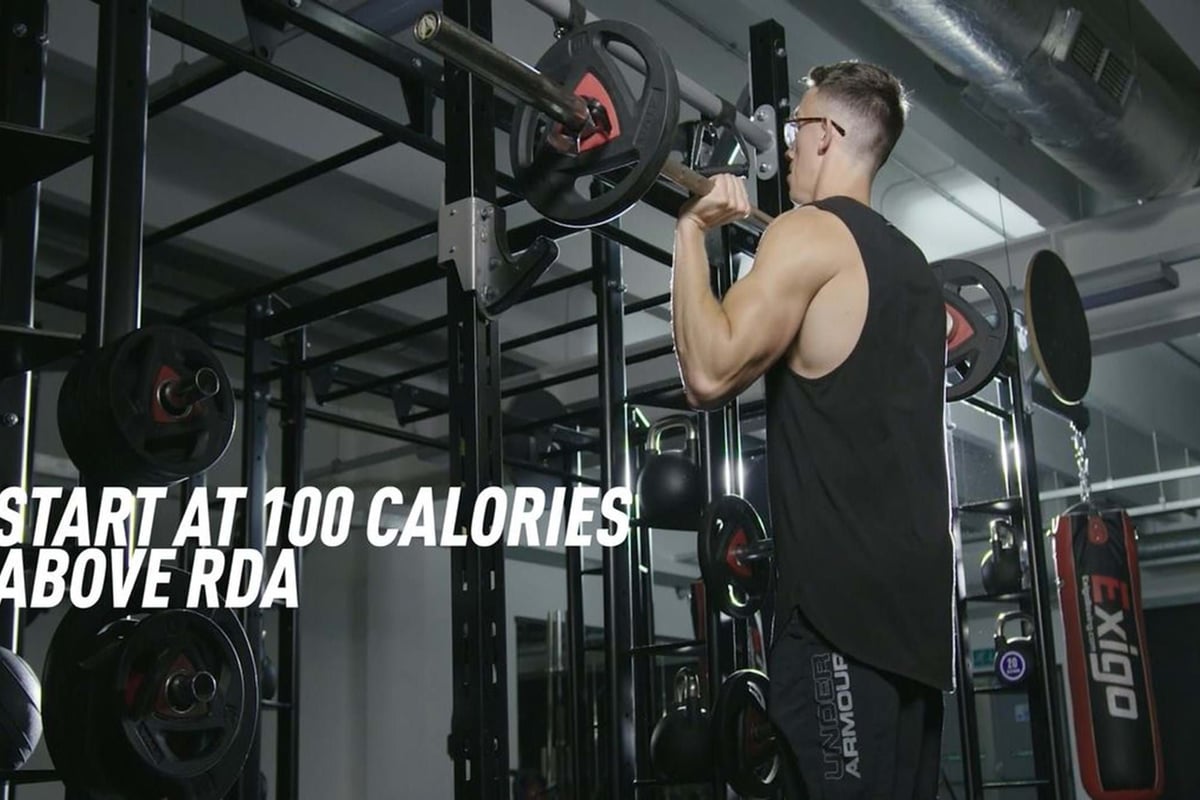 Man lifting barbell weights in gym with text start at 100 calories above RDA