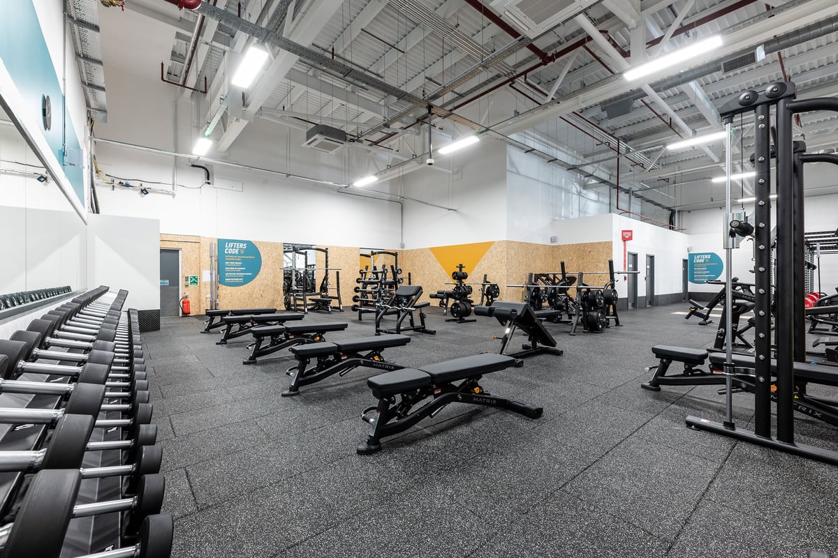 PureGym Rushden: Strength training area with benches, squat racks, free weights, and rubberized flooring.