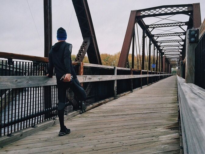 Person in Sportkleidung dehnt Bein auf Holzbrücke mit Metallträgern an bewölktem Tag