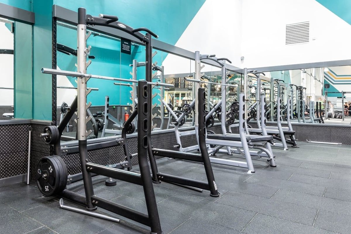 PureGym Aintree: Rows of squat racks, barbells, weight plates, and rubberized flooring in a teal-colored gym.