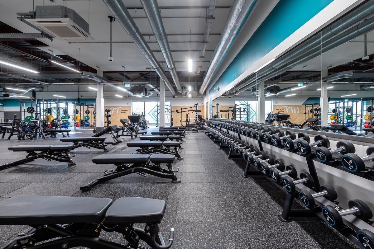 PureGym Craigavon: Rows of weight benches, free weights, and floor mats in a dimly lit weightlifting area.