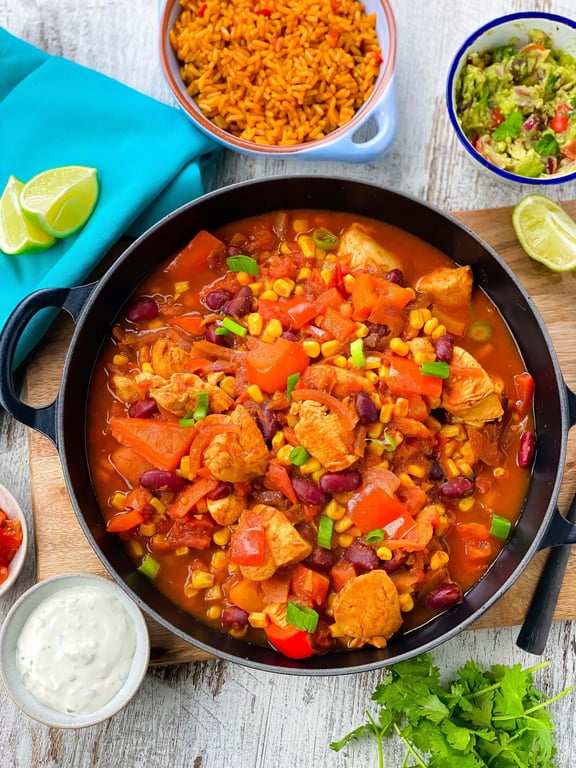 Chicken stew with corn, kidney beans, and red peppers in a black pan with rice and guacamole