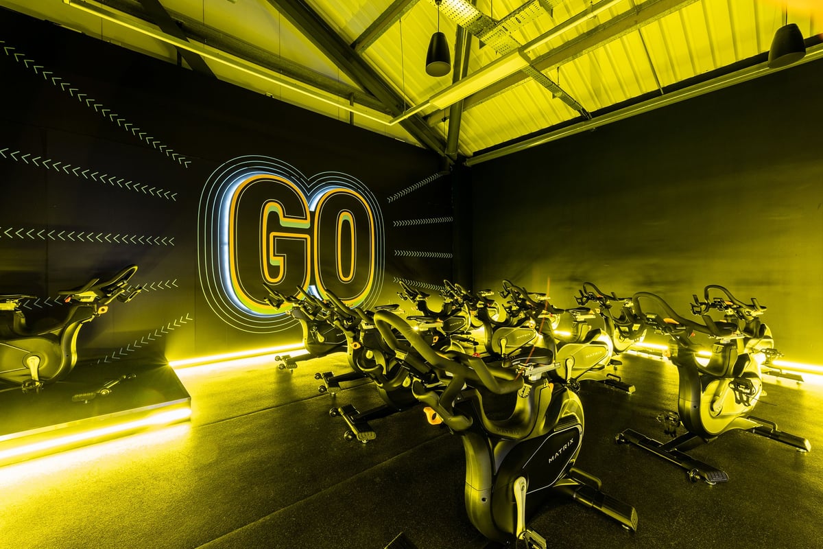 PureGym Chatham: Rows of stationary bikes illuminated by neon "GO" lighting and yellow overhead lights.