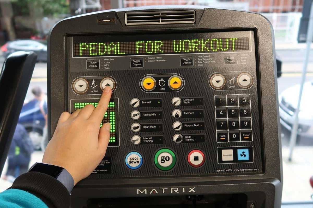 Hand pressing incline button on exercise bike console with PEDAL FOR WORKOUT text above