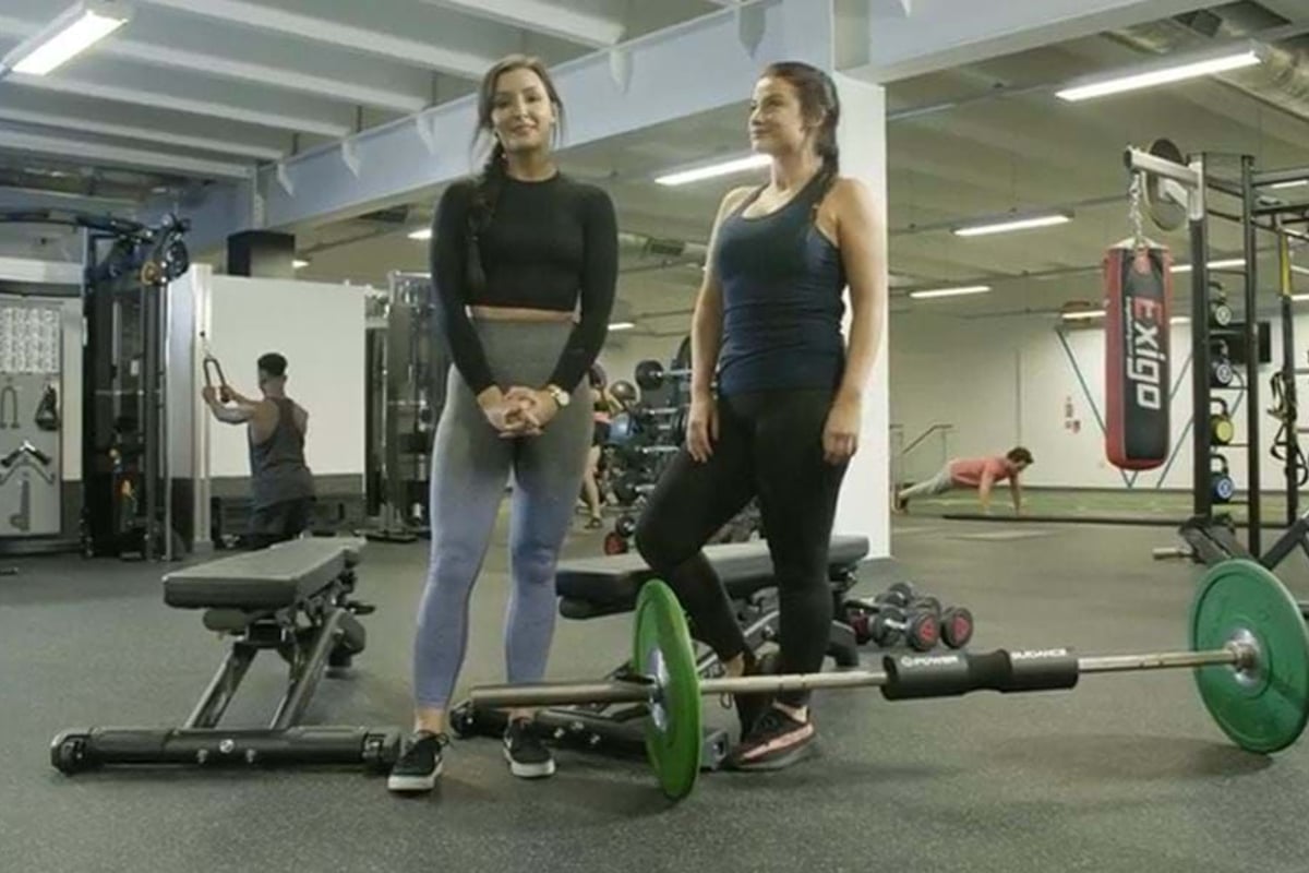 Two women in gym wear standing near a barbell with green weights in a gym.