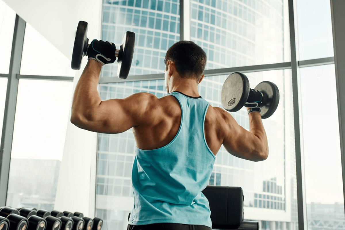 Mann führt Hantel-Schulterpresse in hochgelegenem Fitnessstudio mit Stadtaussicht durch.