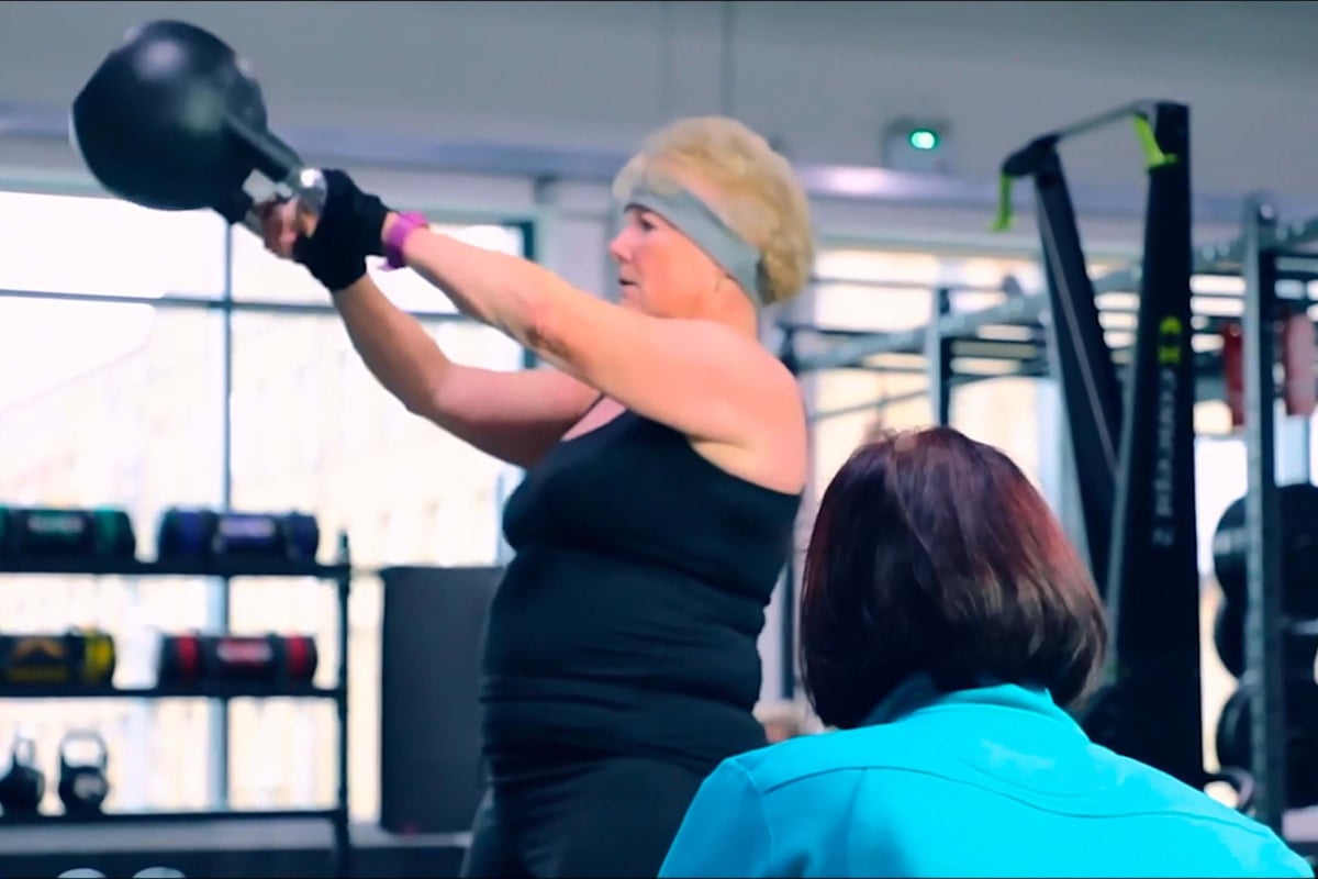 Woman lifting kettlebell while another person watches in gym with equipment in background