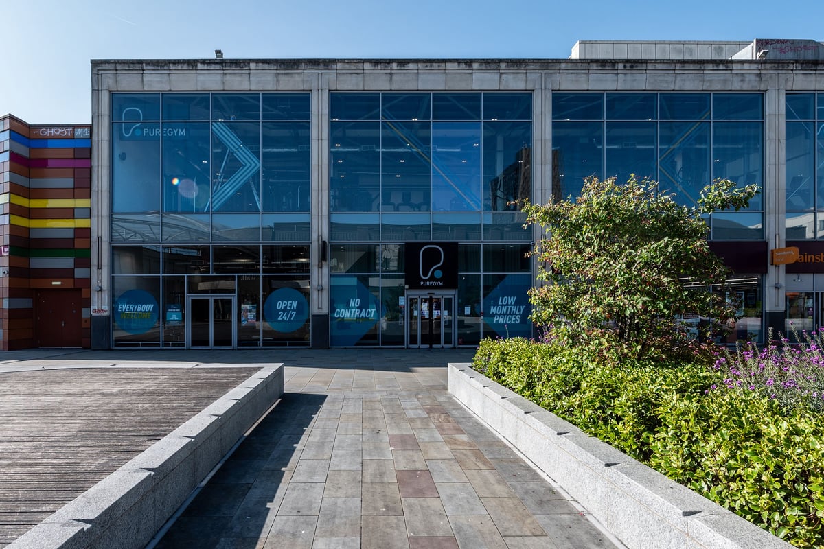 PureGym Newport Wales: Glass-fronted gym building exterior with colourful branding, paved area with planting beds.