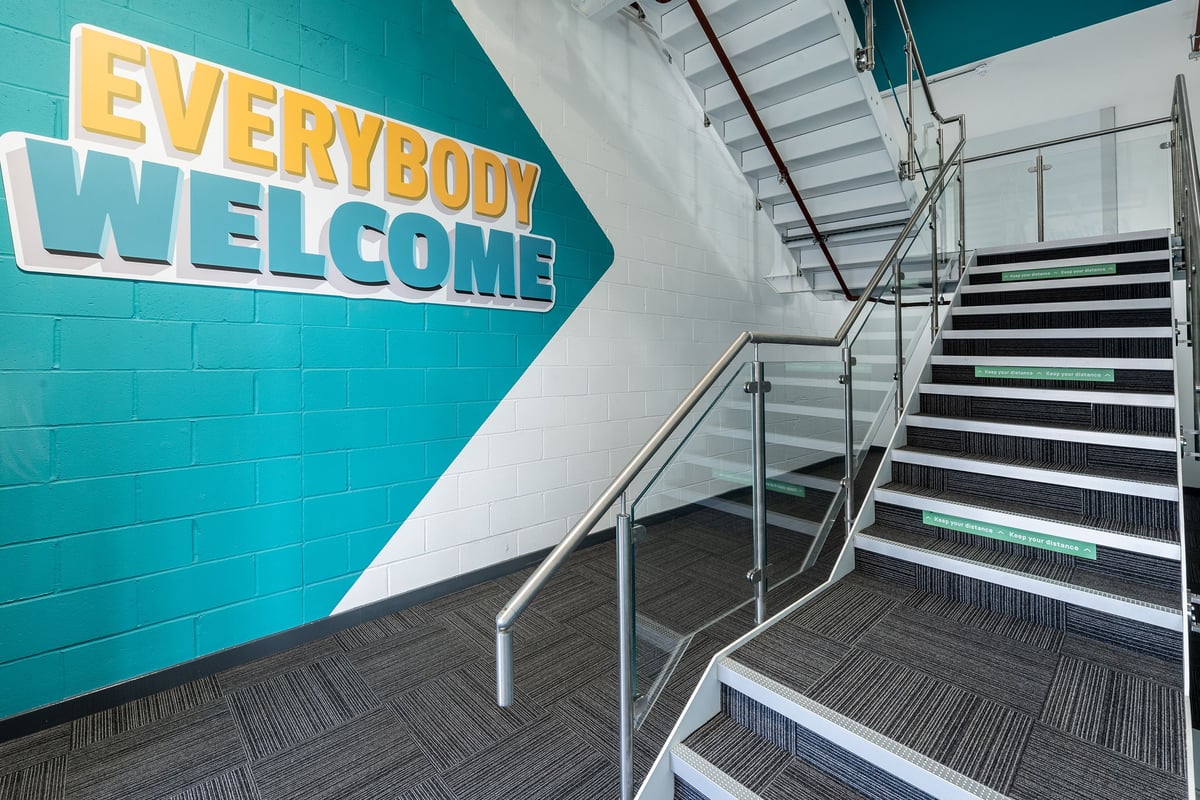 PureGym Maidstone The Mall: Staircase, "Everybody Welcome" sign in teal and yellow, industrial interior design.