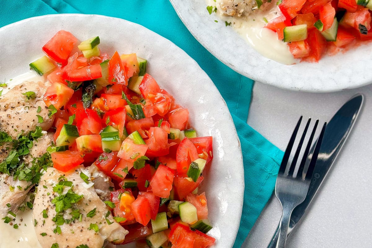 Grilled chicken with creamy sauce and tomato cucumber salad on white plates with cutlery.