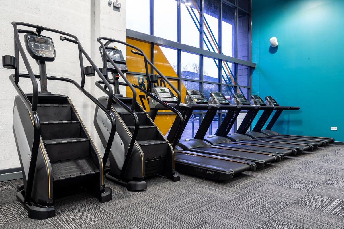PureGym Dunfermline: Rows of treadmills and stair climbers in a modern gym setting with teal walls.